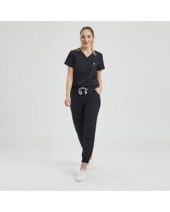 Female model wears a black scrubs set featuring a v-neck top and jogging bottoms with elasticated cuffs and waist drawstring cord. Model's hand is resting in one pocket.
